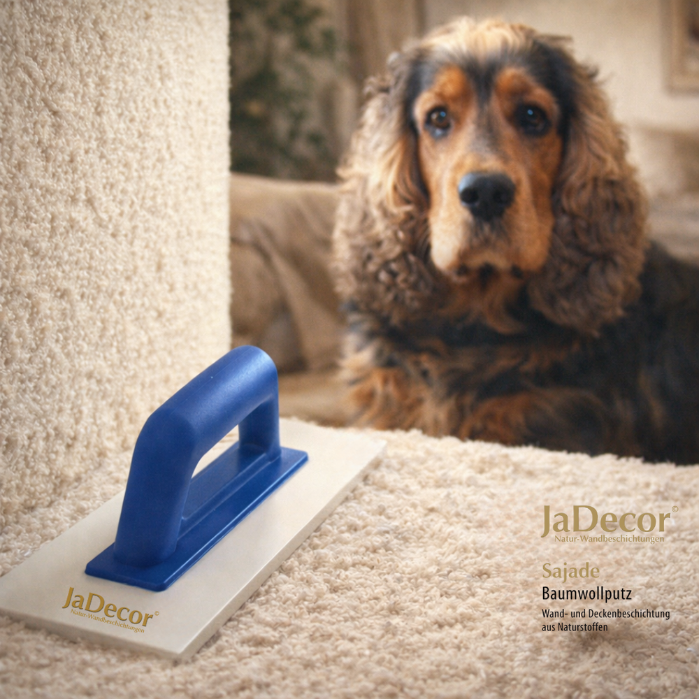 Detailaufnahme von Sajade Baumwollputz mit sichtbarer Struktur, JaDecor Glättkelle im Vordergrund und ruhigem Hund im Hintergrund – natürliche Wandbeschichtung aus Naturstoffen.